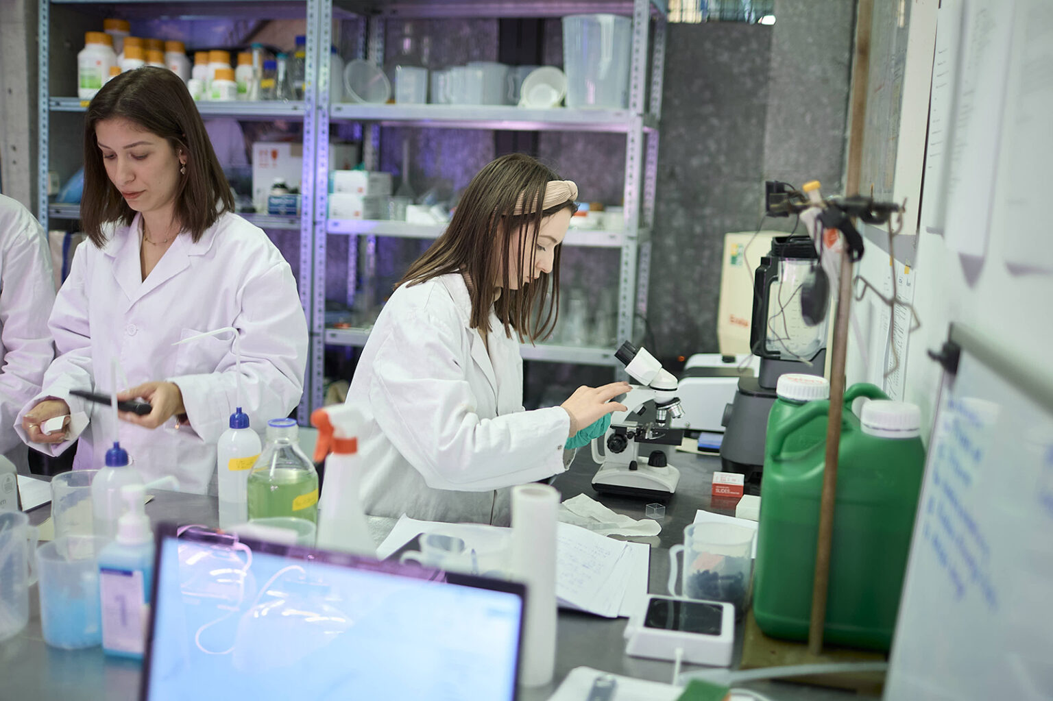 Zwei Personen im Labor, die weiße Laborkittel tragen und mit chemischen Proben sowie einem Mikroskop arbeiten.