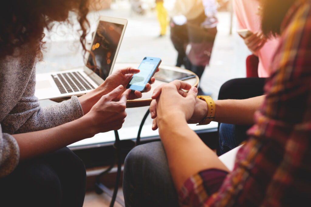 Personen schauen auf ein Smartphone, im Hintergrund steht ein Laptop