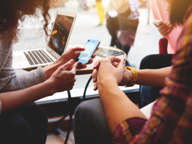 Personen schauen auf ein Smartphone, im Hintergrund steht ein  Laptop