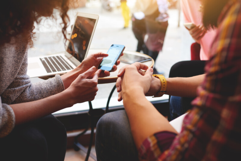 Personen schauen auf ein Smartphone, im Hintergrund steht ein  Laptop