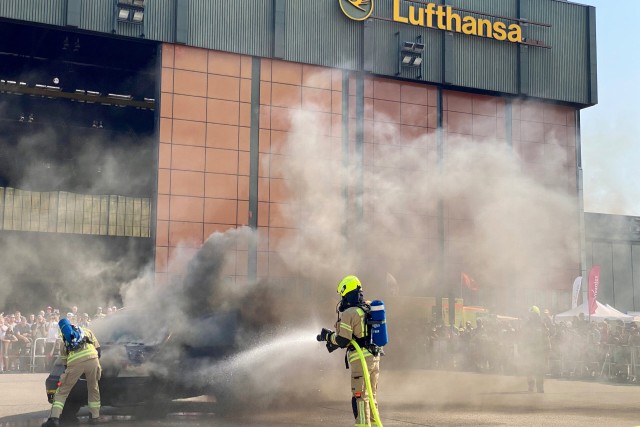 Feuerwehrleute löschen ein brennendes, stark rauchendes Auto vor dem Lufthansa Flugzeughangar.