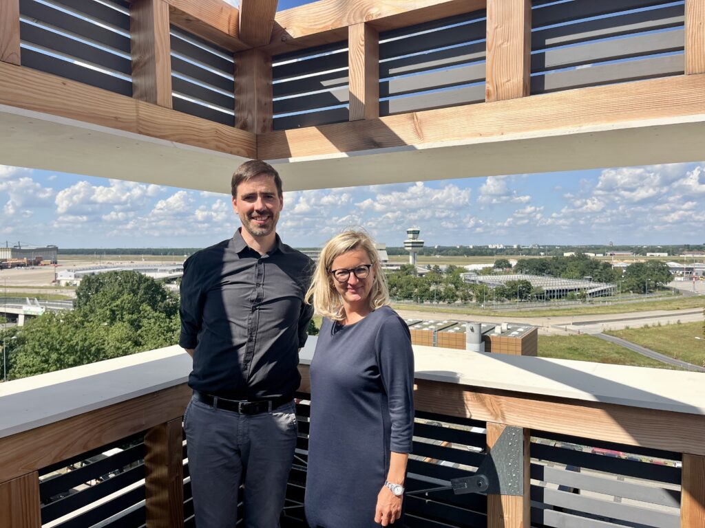 Martina Klement und Dr. Stefan Höffken stehen auf einer Terrasse. Im Hintergrund sieht man das alte Flughafengebäude.