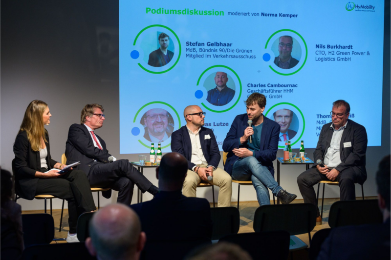 Auf einem Podium hören drei Männer und eine Frau einem Mann mit Mikrofon zu. Im Hintergrund ist eine Präsentation zu sehen