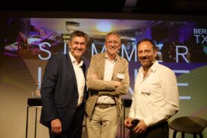 Frank Wolters, Stephan Schütz, und Thomas Willemeit stehen vor einem großen Screen, auf dem Berlin TXL Summer Lounge steht, und lächeln in die Kamera.