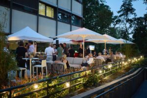 Gäste auf der Terrasse der Berlin TXL Summer Lounge sitzen auf Sofa-Bänken unter Sonnenschirmen in der Dämmerung. Im Hintergrund wird die Podiumsdiskussion übertragen.