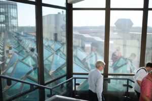 Gäste laufen die Treppe der lichtdurchfluteten, glasüberdachten Haupthalle des Terminals hinunter. Dabei sieht man im Hintergrund das Flughafengelände.