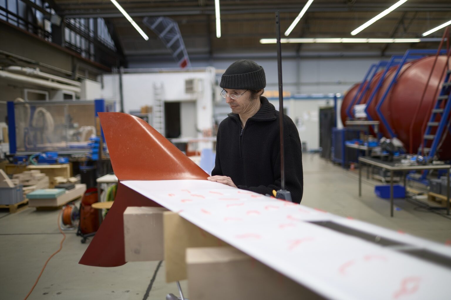 Eine Person in schwarzer Kleidung arbeitet konzentriert an einem orange-weißen Windkraftflügel in einer Produktionshalle