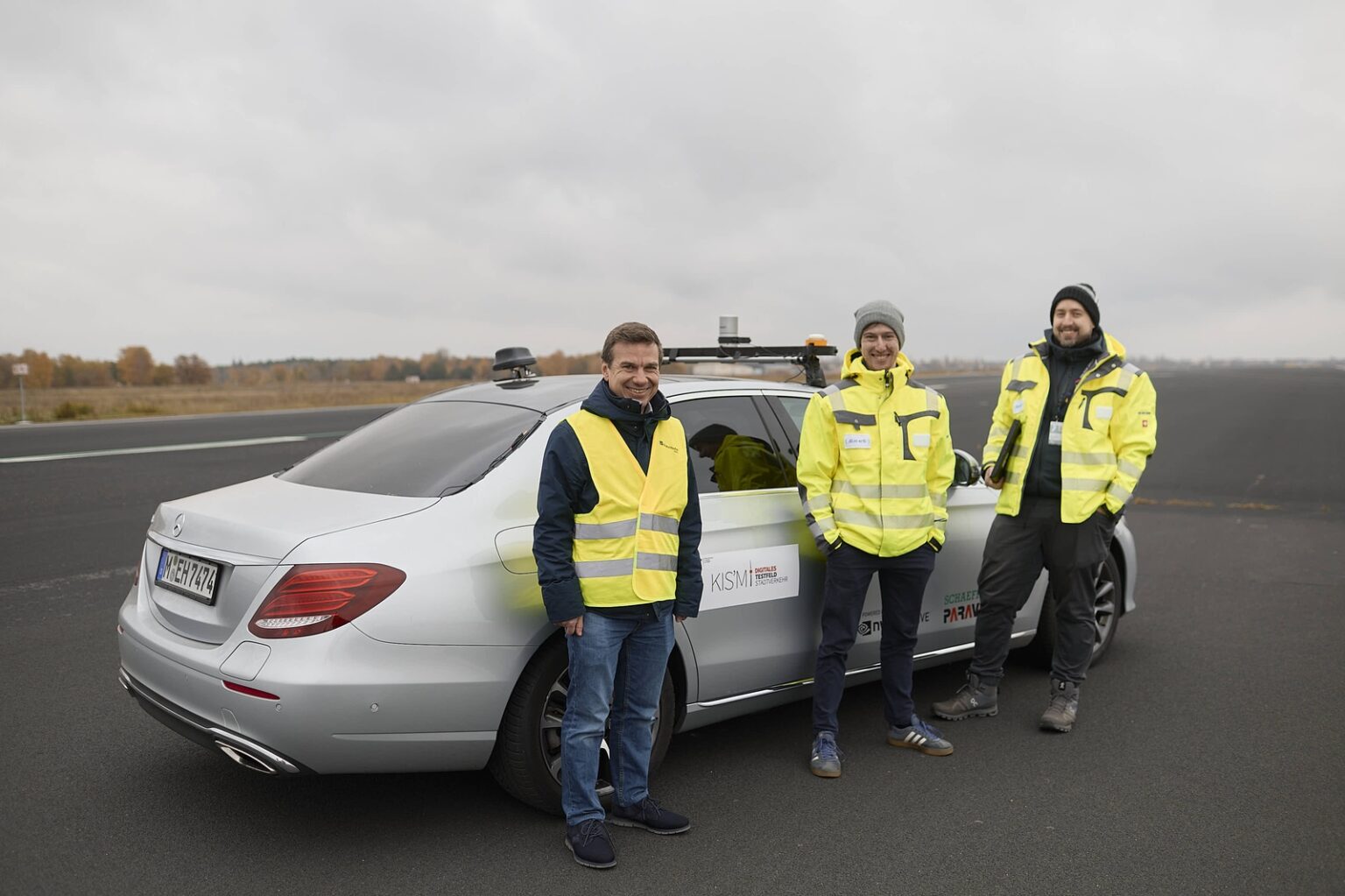 Drei Personen in Sicherheitswesten stehen vor einem autonomen Fahrzeug auf einem Flugplatz.