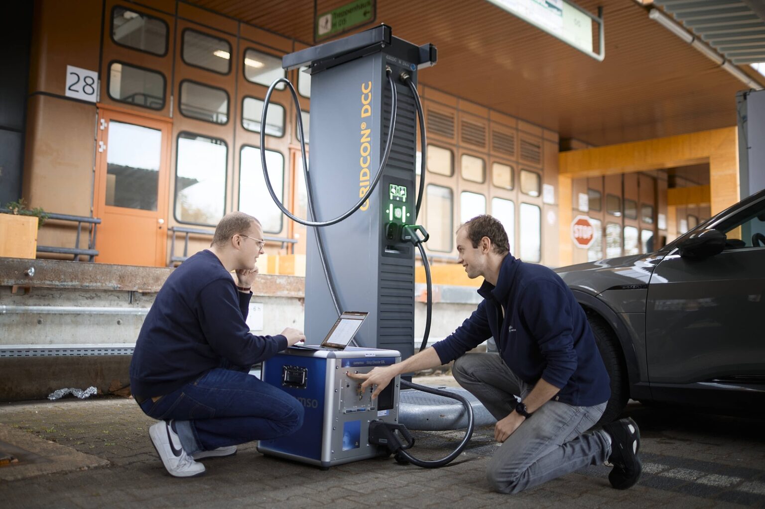 Zwei Fachkräfte arbeiten an einer Ladesäule für Elektrofahrzeuge auf einem Werksgelände.