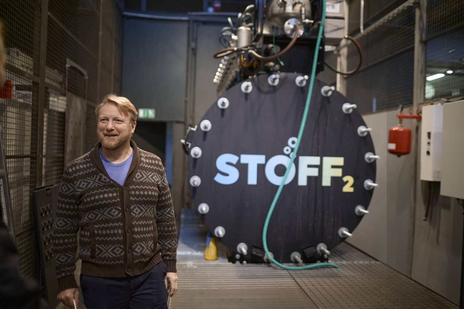 Eine Person in braunem Pullover steht vor einer großen Wasserstoffanlage mit der Aufschrift "STOFF2".